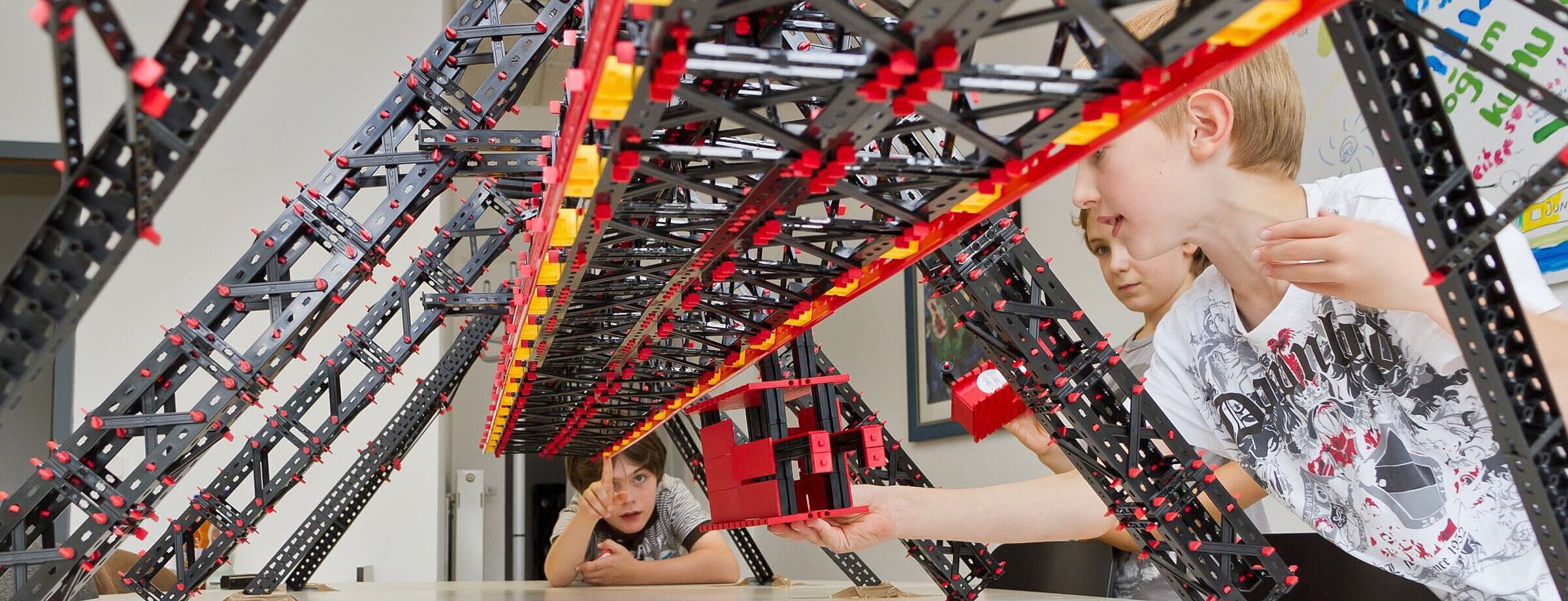 Drei Kinder spielen mit einem Modellbau der Schwebebahn von Fischertechnik. 