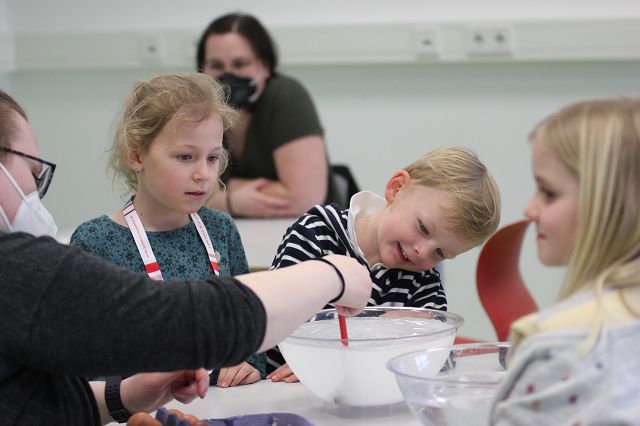 Kinder im Kurs "Zaubereien aus dem Kühlschrank" beugen sich über eine Schüssel mit Flüssigkeit, die von der Dozentin gerührt wird. 